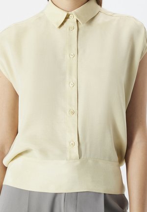 Blouse jaune clair à manches courtes avec col et six boutons devant, associée à un pantalon gris, visible du cou aux hanches.