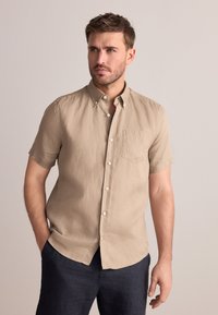 Kurzärmeliges Hemd aus leichtem beigem Leinen mit Knopfleiste, Vorder- tasche und legerer Passform. Kombiniert mit dunkelblauen Hosen.