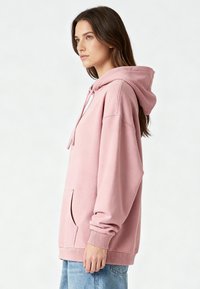 Felpa rosa con cappuccio dal taglio ampio, tasca frontale, polsini a coste e texture morbida. Il modello indossa jeans azzurri.