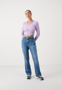 Top longo de manga curta em lilás com textura de canelado, combinado com jeans de pernas retas azuis desbotados e tênis pretos e beges. Colar de corrente simples.