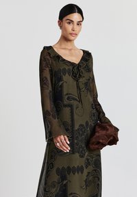 Robe longue vert olive avec des motifs paisley noirs, encolure festonnée et manches transparentes. Le mannequin tient un tissu marron dans sa main.