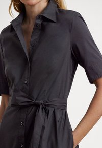 Camisa preta de manga curta feita de um tecido liso, com frente abotoada, gola e cinto à cintura atado.