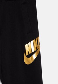 Juodos sportinės kelnės su blizgia auksine Nike logotipu ir šluotele, su šonine kišene funkcionalumui, pateiktos prieš neutralų foną.