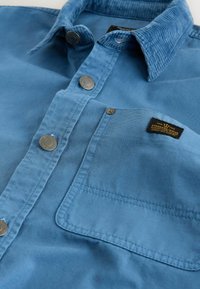 Blå skjorte med knapper lavet af bomuld. Har en fløjlskrave, metaltryklåse og en brystlomme med et brand mærke. Blød tekstur.
