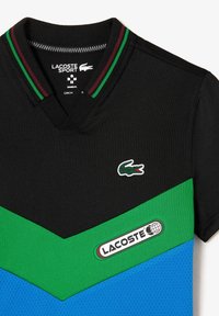 Black short-sleeve polo shirt featuring a green and blue chevron design. Ribbed collar in red, green, and black. Lacoste logo on chest.