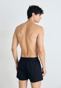 Schwarze Baumwollshorts mit einem elastischen Bund, die eine lockere Passform bieten. Das Modell ist von hinten zu sehen und zeigt eine glatte Textur.