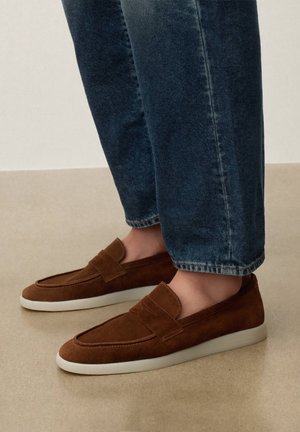 Barna szarvasbőr könnyen felvehető mokaszinek fehér talppal, kék farmernadrággal viselve, bézs padlón.