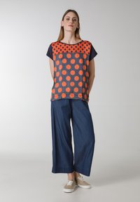 Maglietta colorata con pois arancioni e blu navy, scollatura rotonda, maniche corte; abbinata a pantaloni in denim blu a gamba larga e sneakers color sabbia.