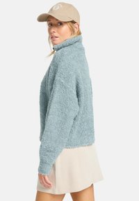 Maglione azzurro chiaro, morbido, con colletto alto e maniche lunghe, abbinato a un berretto beige. Lo sfondo è bianco semplice, concentrandosi sull'abbigliamento.