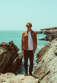 Veste en velours côtelé marron sur une chemise blanche, pantalon sombre, se tenant sur une rive rocheuse avec l'océan en arrière-plan. Ciel clair et lumière du soleil.