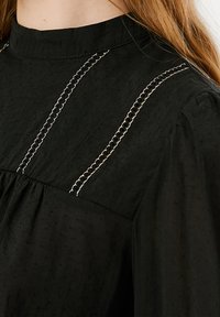 Schwarze Bluse mit hohem Kragen, strukturiertem Stoff und dekorativen weißen und schwarzen Nähten an den Schultern.