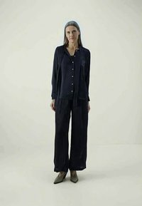 Blusa in satin blu navy con chiusura a bottoni e maniche lunghe, abbinata a pantaloni blu navy a gamba larga e tacchi a punta. Texture liscia.