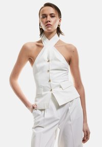 Witte haltertop met een aansluitende bodice, voorzien van een rij gouden knopen en zijzakken. Gecombineerd met hoge, witte broeken.