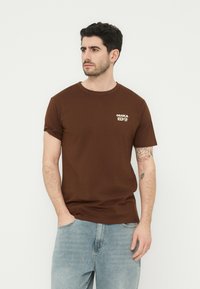 Άνδρας με σκούρα μαλλιά, που φοράει καφέ μπλούζα με την ένδειξη "Osaka" και ανοιχτόχρωμο μπλε τζιν, στέκεται με το ένα χέρι στην τσέπη, κοιτώντας προς τα αριστερά.