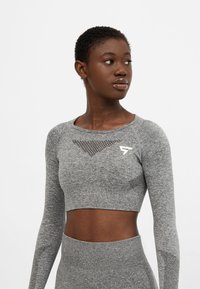 Šedý cropped top s dlhým rukávom a sieťovými panelmi, priliehavý strih a logo akcent. Mäkka textúra, vhodný na aktívne nosenie.