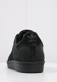 Svarta Adidas-sneakers sedda bakifrån, med en hälla i mocka med Adidas-logotyp, texturerad sula och ovandel i mocka.