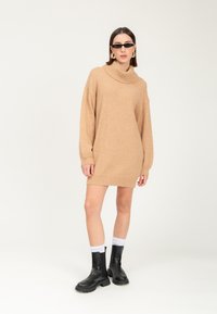 Tan strikket sweaterkjole med høj krave, lange ærmer og ribbede manchetter, parret med sorte ankelstøvler og hvide sokker.