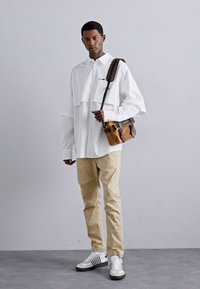 Cămașă oversized albă cu design stratificat, pantaloni bej și rucsac maro. Încălțămintea are dungi albe și negre. Textura țesăturii este netedă.