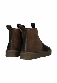 Hnědé semišové a kožené kotníkové boty s gumovou podrážkou, zlatým zipem a poutky pro snadné obouvání. Branding viditelný na patě.