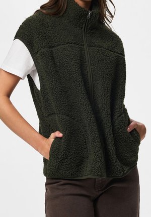 Personne portant une veste sans manches en polaire texturée vert foncé zippée sur un t-shirt blanc à manches courtes, les mains dans les poches de la veste.