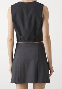 Maßgeschneiderte dunkelgraue cropped Weste mit geradem Ausschnitt und Rückenverschlussdetail, kombiniert mit einem plissierten Rock aus passendem Stoff.