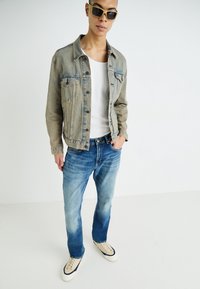Ljusblå denimjacka med en lätt nött finish, vit T-shirt, urtvättade blå jeans och beige sneakers med marinblå detaljer.