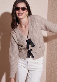 Beige cable-knit cardigan with a V-neck, belted by black ribbon ties, featuring two pockets and ribbed sleeve cuffs, worn over white jeans.