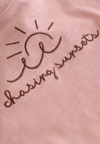 Broderie marron d’un lever de soleil et texte "chasing sunsets" sur tissu rose doux, montrant la texture et les détails des coutures.