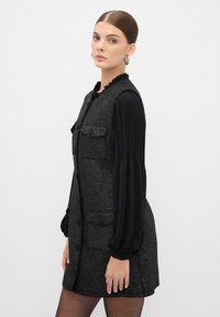 Chaleco negro texturizado con dos bolsillos frontales, cierre de botones y una blusa negra de manga larga con pliegues debajo.