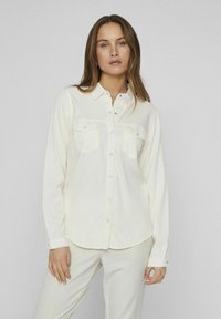 Camicia bianca a maniche lunghe in tessuto liscio, con due tasche sul petto e una chiusura con bottoni sul davanti. Orlo arrotondato e polsini con bottoni.