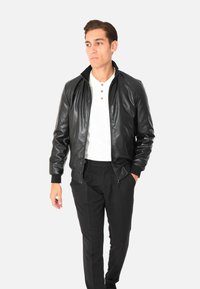 Giubbotto bomber in pelle nera con cerniera frontale, polsini e colletto a costine, abbinato a una maglietta henley bianca e pantaloni neri.