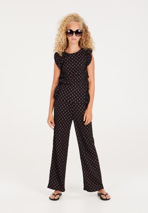 Zwarte jumpsuit met gerimpelde mouwen en een rood bloemenpatroon. Wijde broekstijl gecombineerd met zwarte slippers en oversized zonnebril.