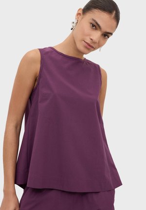 Femme portant un haut violet sans manches et un pantalon violet assorti, avec les cheveux tirés en arrière et des boucles d'oreilles argentées imposantes.