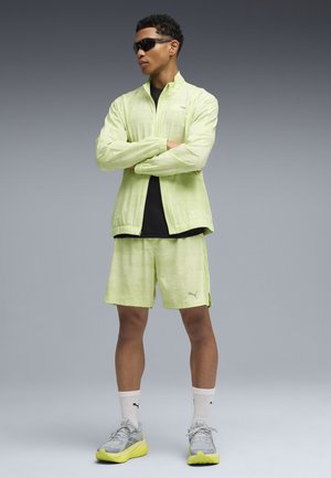 Limoengroene sportjas en korte broek met een gestructureerde afwerking, gecombineerd met grijze sneakers met neonaccenten. Zwart T-shirt en zonnebril.