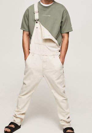 Homme portant une salopette beige avec une bretelle défait, un t-shirt vert à manches courtes et des sandales noires à enfiler, debout les mains dans les poches.