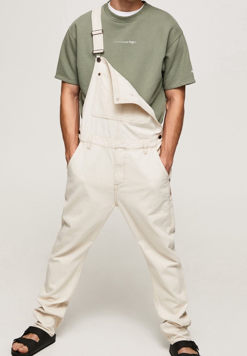 Homme portant une salopette beige avec une bretelle défait, un t-shirt vert à manches courtes et des sandales noires à enfiler, debout les mains dans les poches.