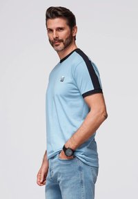 Světlomodré tričko s černými akcentními rukávy, kulatým výstřihem, s detailem loga vpředu, doplněné světle modrými džínami a černými hodinkami.