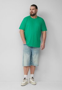 Zielona koszulka z krótkim rękawem w luźnym kroju, w połączeniu z jasnoniebieskimi szortami dżinsowymi. Model ma na sobie białe trampki z szarymi akcentami oraz białe skarpetki.