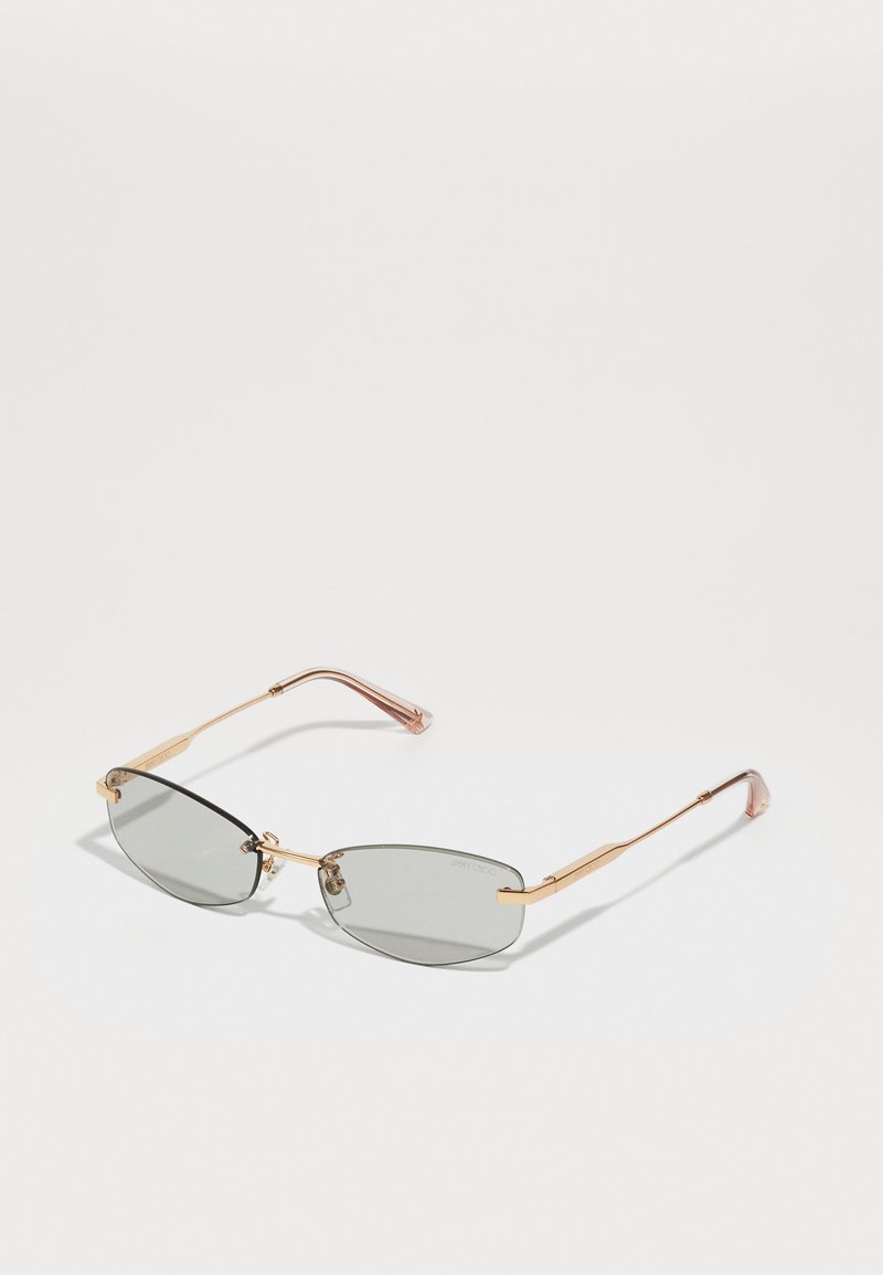 Óculos de sol com lentes cinza escurecidas, armação fina em metal dourado, forma alongada e pontas das hastes acentuadas em rosa. Design minimalista com uma marcação subtil.