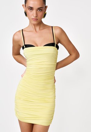 Kobieta z zaczesanymi do tyłu włosami, ubrana w marszczoną żółtą mini sukienkę na czarnym biustonoszu, pozująca z rękami za plecami na jednolitym białym tle.