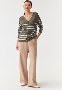 Olivgrüner Cardigan mit weißen horizontalen Streifen, V-Ausschnitt, sieben Knöpfen und gerippten Bündchen, kombiniert mit beigen, weiten Hosen.