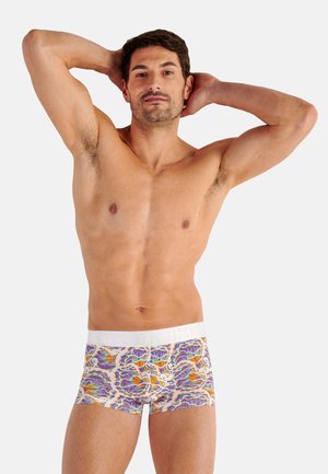 Mann mit kurzen dunklen Haaren steht oben ohne mit den Händen hinter dem Kopf und trägt farbenfrohe Boxershorts mit Blumenmuster vor hellem Hintergrund.