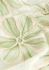 Zbliżenie tkaniny z wypukłą, fakturowaną wyszywanką tworzącą zielone kwiatowe wzory obrysowane kremowym kolorem na jasnym tle.