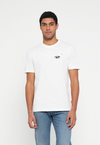 Λευκό βαμβακερό T-shirt με στρογγυλή λαιμόκοψη, που διαθέτει μικρό μαύρο γραφικό τύπωμα ματιού και οριζόντια ρίγα στο στήθος.