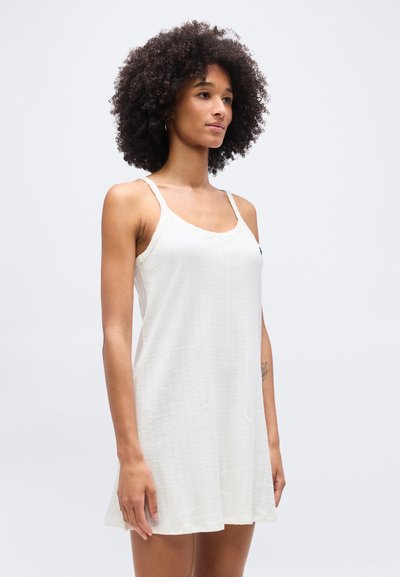 Γυναίκα με σγουρά μαλλιά που φοράει λευκό αμάνικο πλεκτό φόρεμα, όρθια μπροστά σε απλό ανοιχτό γκρι φόντο, κοιτώντας ελαφρώς προς τα δεξιά.