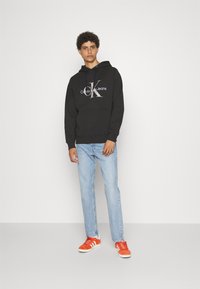 Melnā kapuci ar "Calvin Klein Jeans" logo sudrabainā krāsā, apvienota ar gaiši ziliem džinsiem un oranžām kedām. Modelis stāv uz baltas fona.