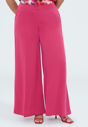 Donna che indossa pantaloni larghi rosa acceso e sandali rossi con tacco alto, in piedi con le mani nelle tasche davanti a uno sfondo semplice.