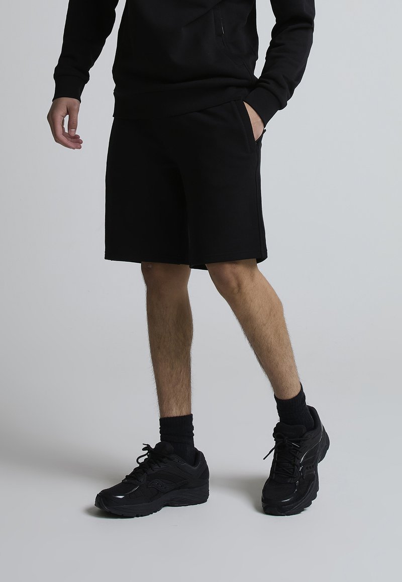 Personne debout portant un short noir, un haut à manches longues noir, des chaussettes noires et des chaussures de sport noires sur un fond gris clair.