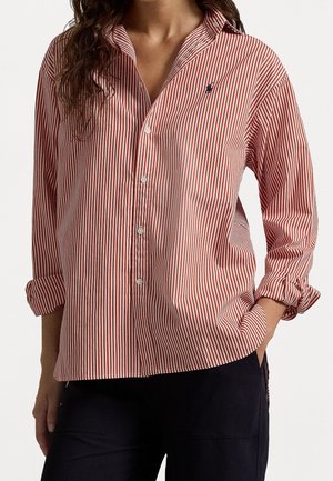 Woman wearing a red and white vertically striped button-up shirt with rolled sleeves and dark pants, hand in pocket on a plain background.