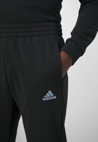 Pantalones de chándal negros con cintura elástica, que presentan un logo de Adidas bordado en gris en la parte inferior de la pierna izquierda. Textura de tela suave.